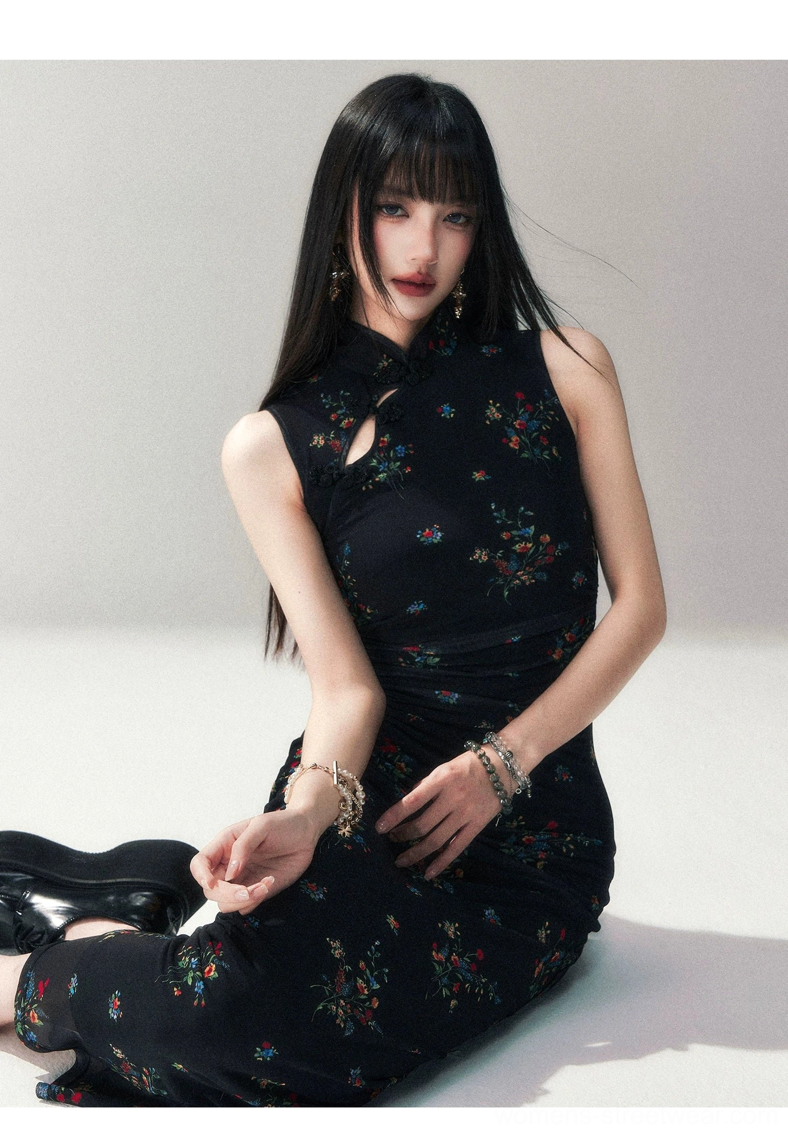 Three-quarter seated view of a black floral sleeveless qipao-inspired dress with Mandarin collar, fitted silhouette, floral pattern, and layered bracelets