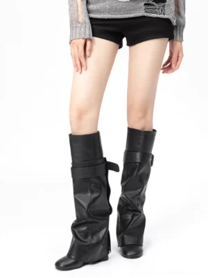 Model wearing black foldover knee-high streetwear boots with draped shaft and buckle strap in a front view