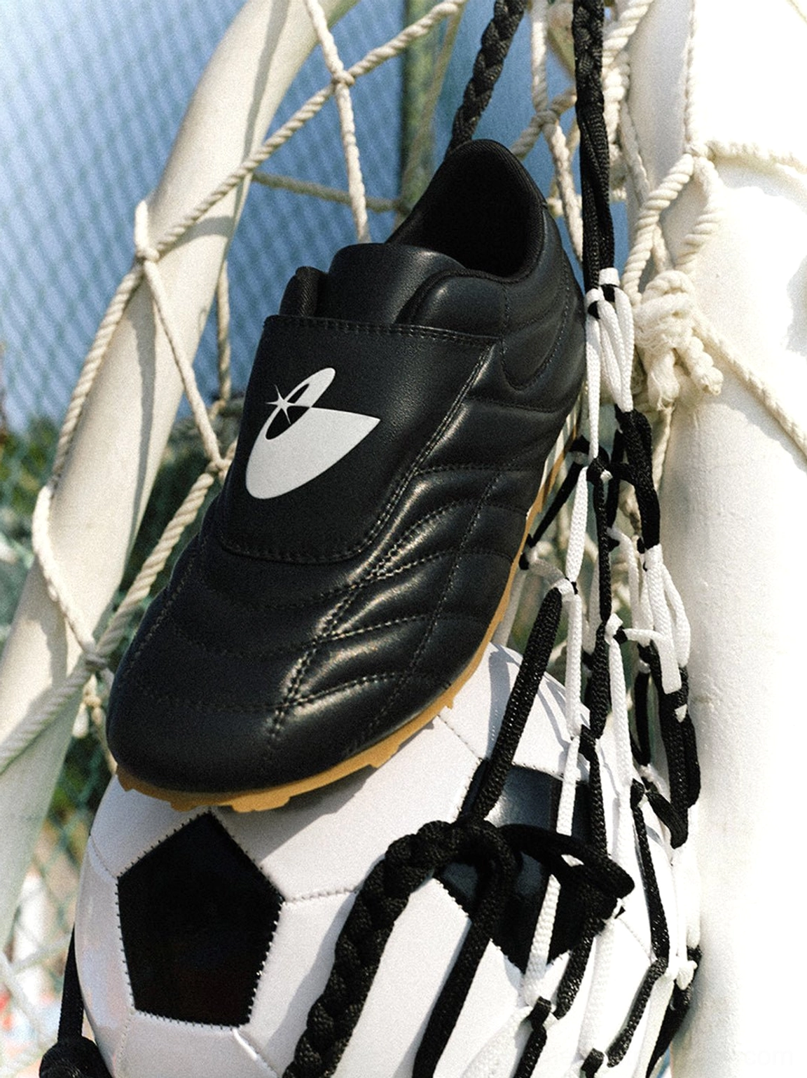 Black football trainer sneaker hanging on a soccer goal net with a football below, showing quilted upper and gum outsole
