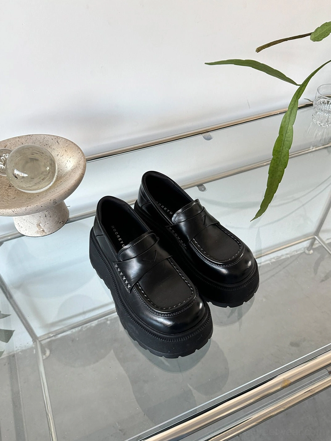 Black glossy rounded-toe chunky platform loafers displayed from two angles on a minimalist glass table
