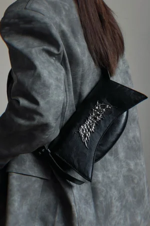 Black gothic metal logo shoulder bag worn at the hip with a distressed grey leather coat in streetwear styling