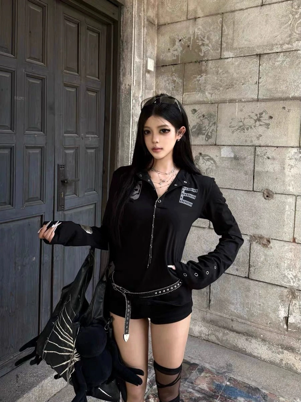 Black gothic zip-front long sleeve top styled with metal zipper details, studded letter decoration, and eyelet cuff accents in an outdoor editorial setting