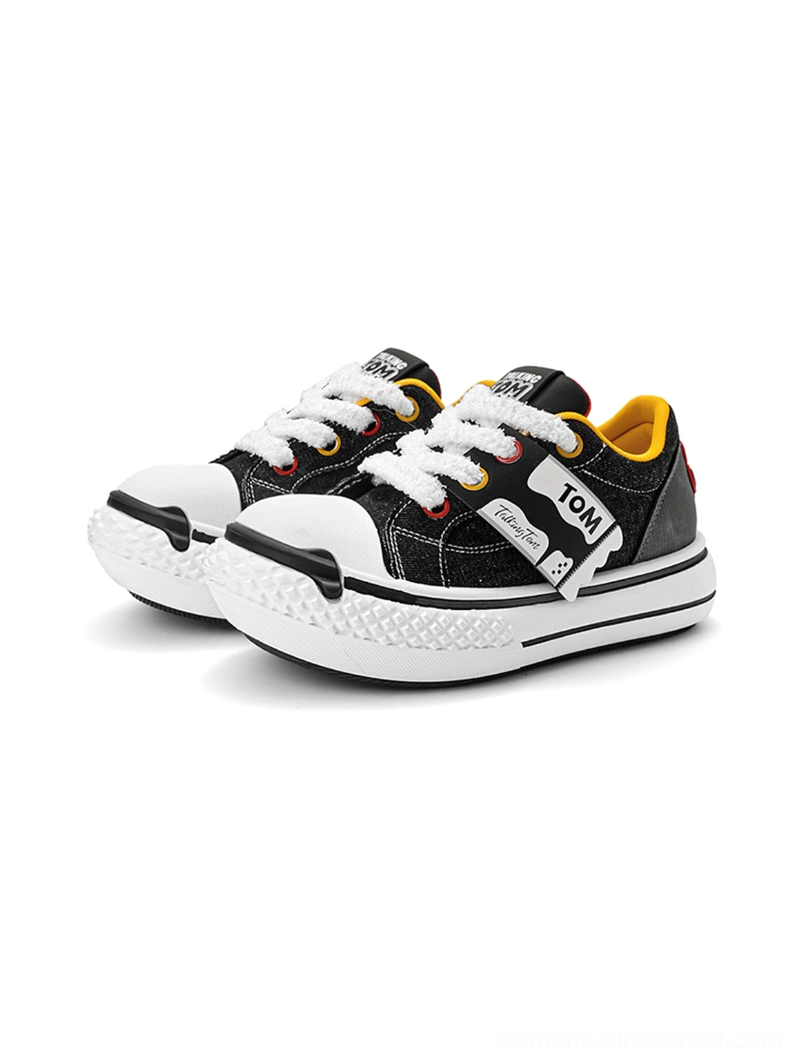 Black graphic low-top sneakers shown from a front three-quarter angle with thick white soles, decorative strap, laces, and yellow lining