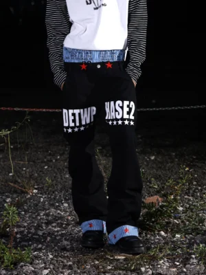 Low-angle front view of black graphic wide-leg jeans featuring red star waist details, blue striped turn-up cuffs, and relaxed streetwear styling