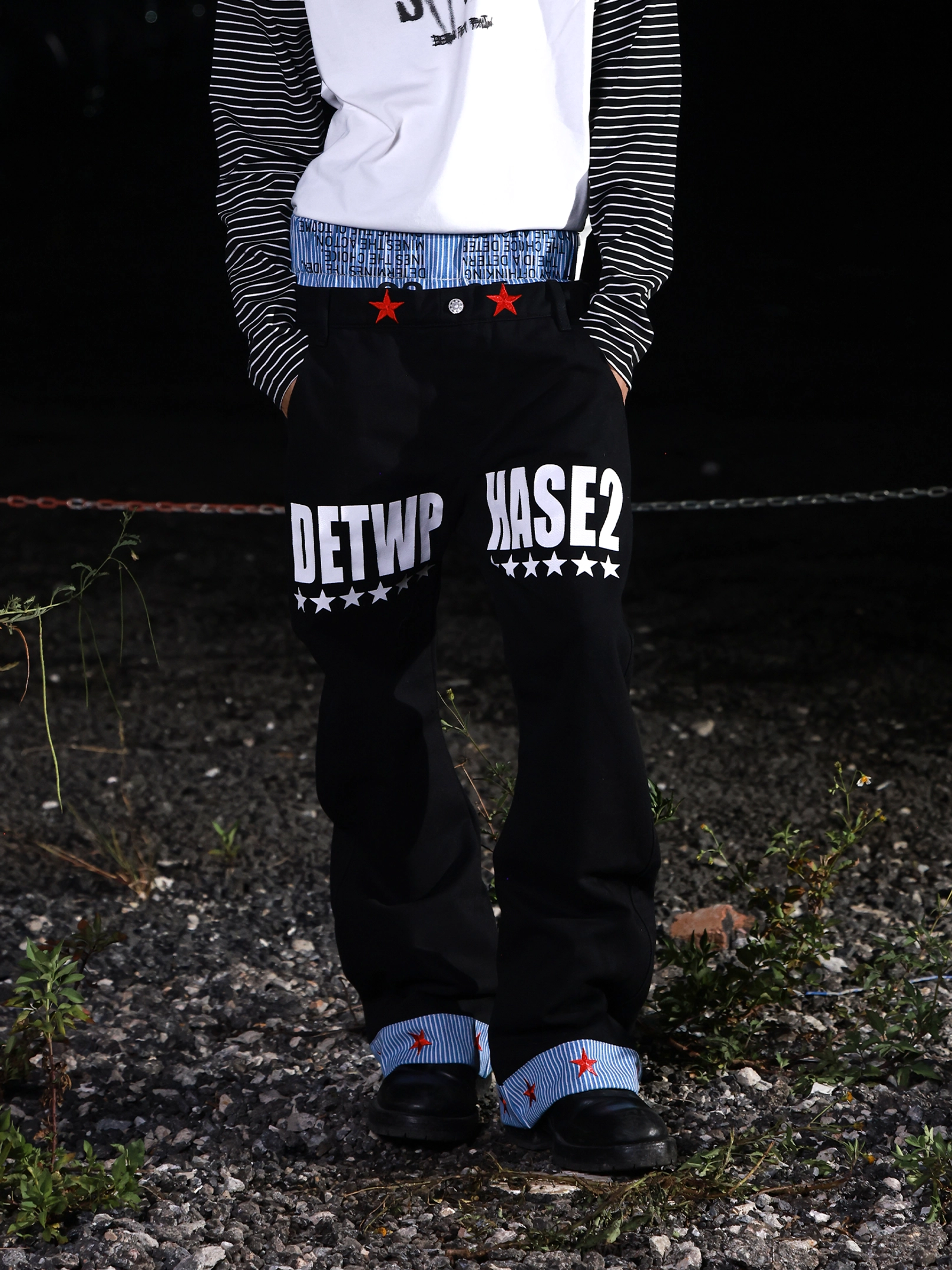 Low-angle front view of black graphic wide-leg jeans featuring red star waist details, blue striped turn-up cuffs, and relaxed streetwear styling