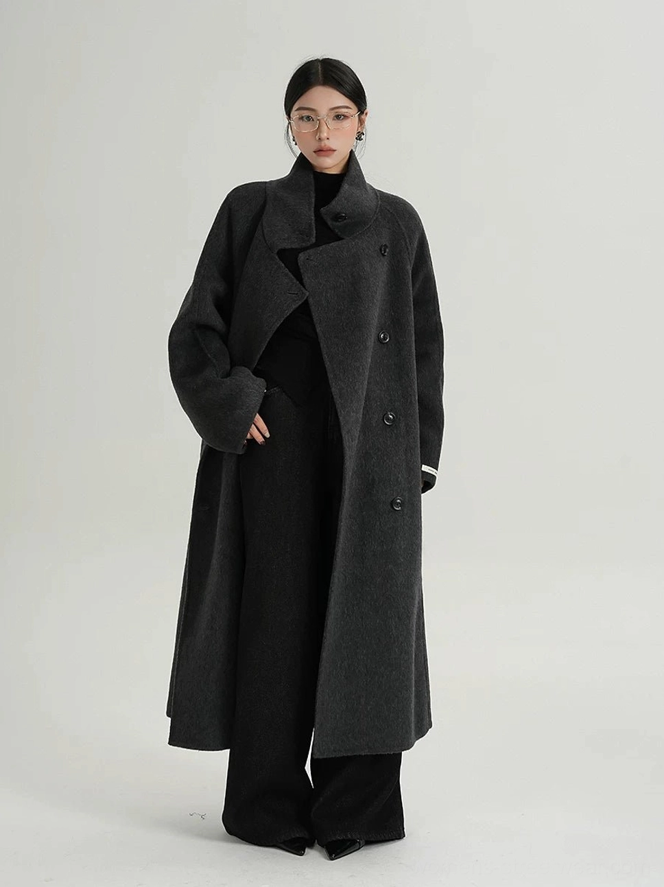 Full-body front view of a woman wearing an oversized dark gray black double-breasted funnel neck wool cocoon coat with dropped sleeves and long streetwear silhouette