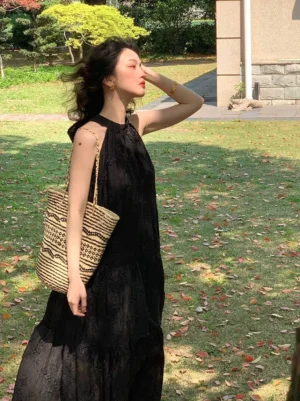 Black halter neck lace maxi dress with tied neckline and floor-length tiered silhouette styled on a park lawn with woven tote bag