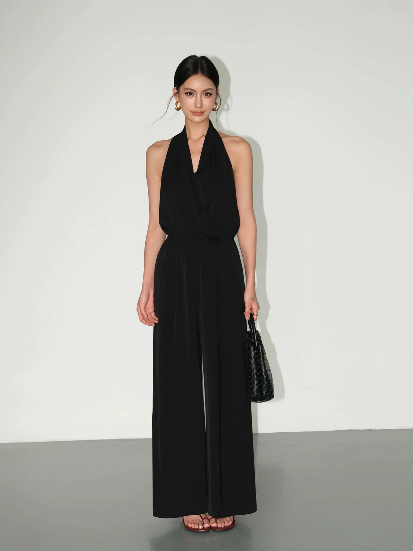 Model wearing a black halter wide-leg jumpsuit with draped wrap neckline and quilted handbag