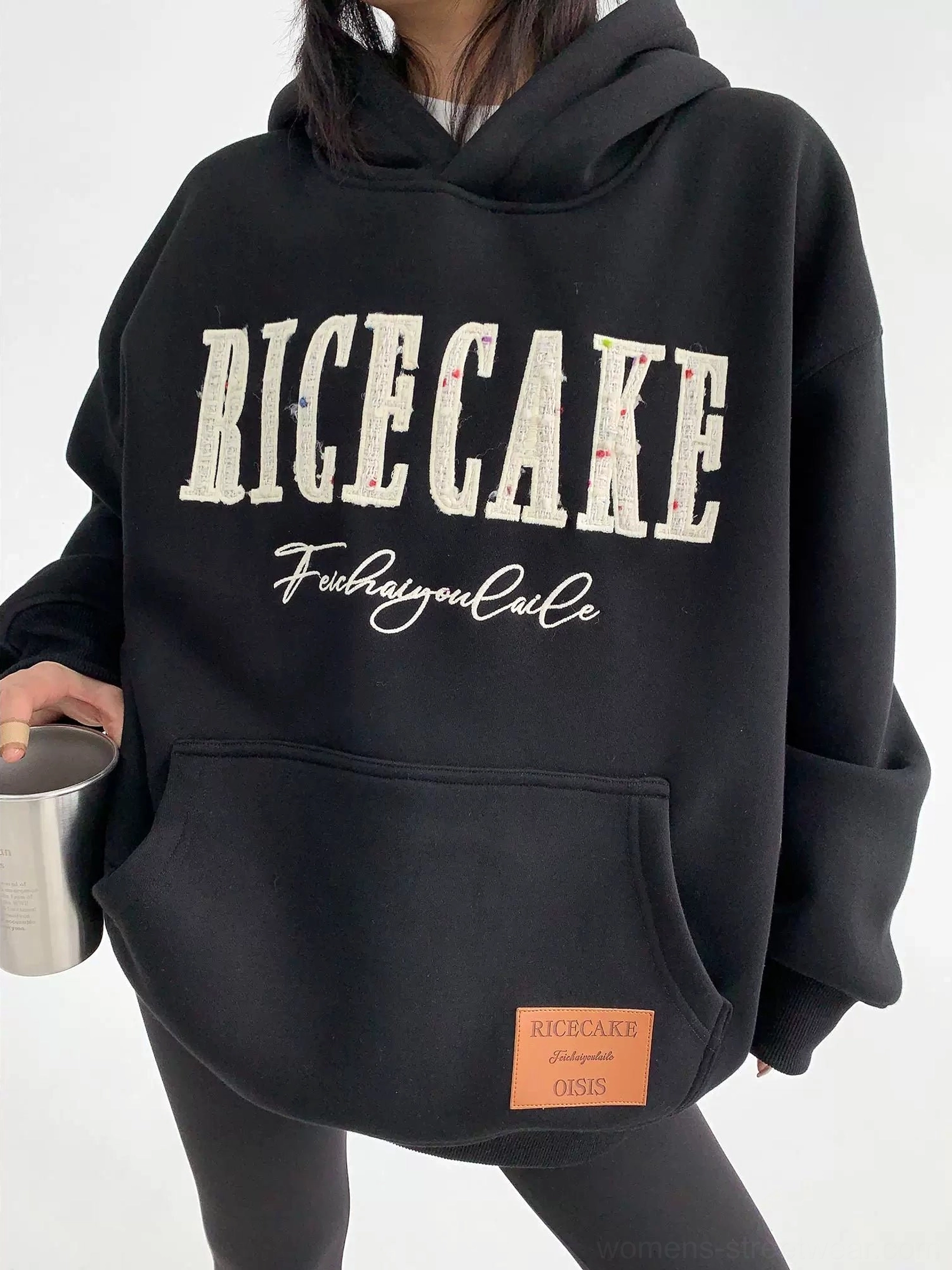 Close-up of black oversized heavyweight fleece hoodie showing layered varsity lettering, kangaroo pocket, and faux-leather hem patch