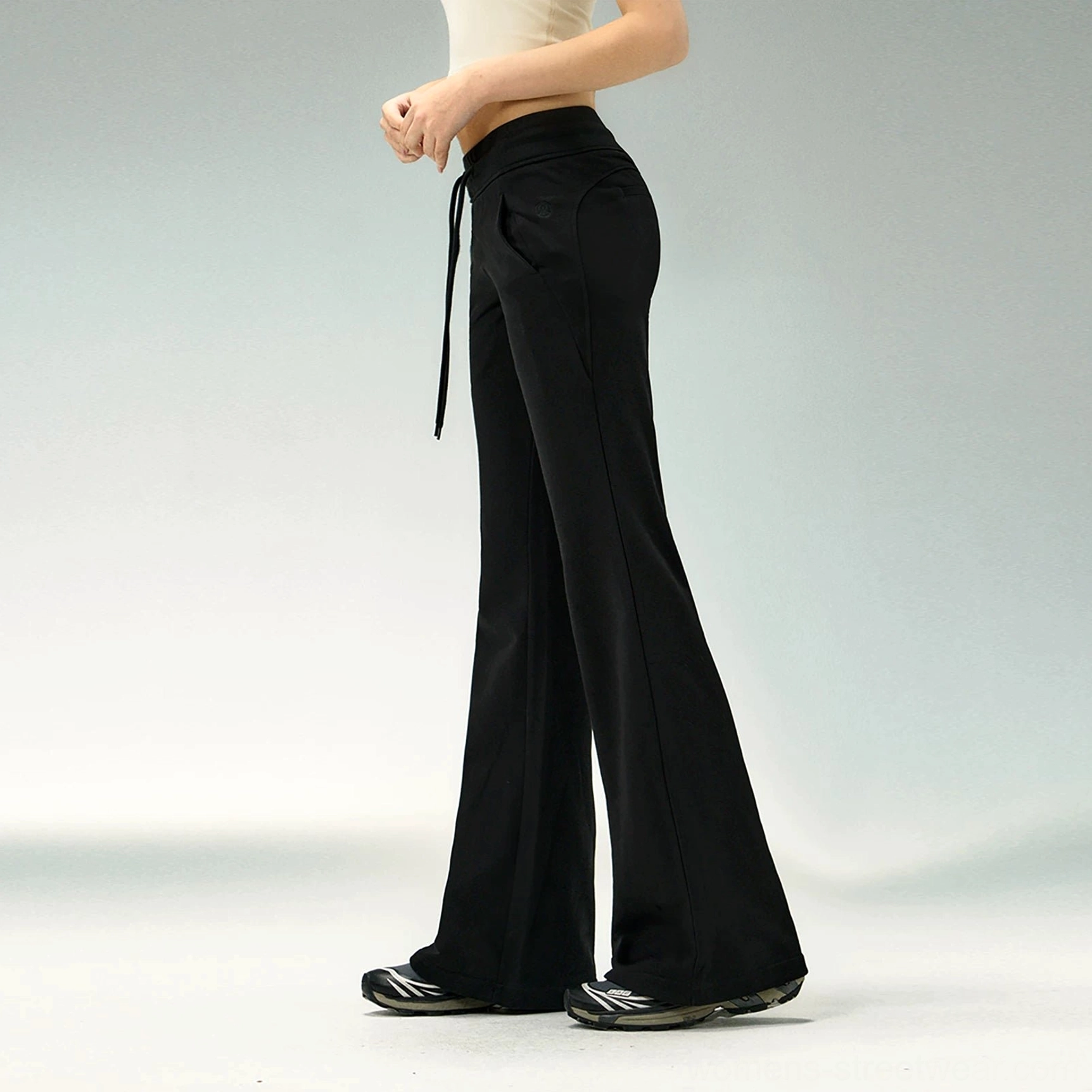 Back side view of black high-waisted flared sweatpants with a sleek smooth finish, elongated silhouette, cropped top, and casual sneakers