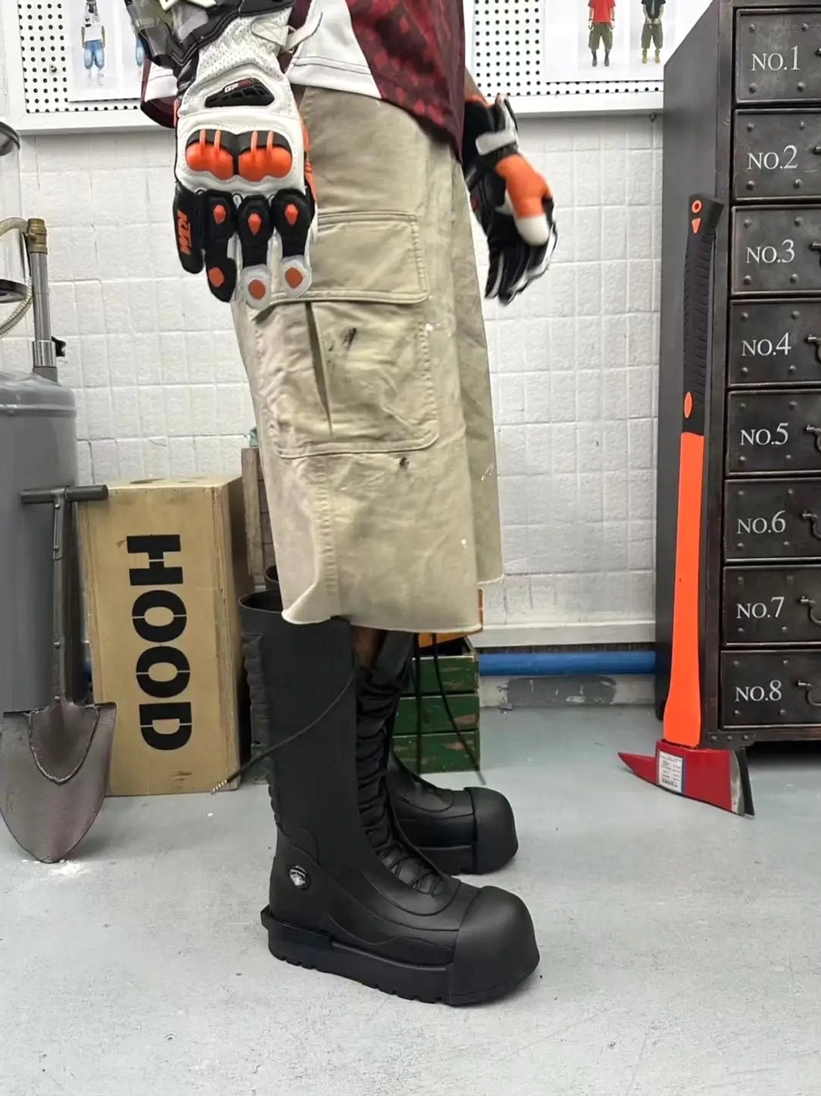 Black knee-high platform combat boot worn with cargo shorts in an industrial studio, showing the tall ribbed shaft and rugged streetwear silhouette