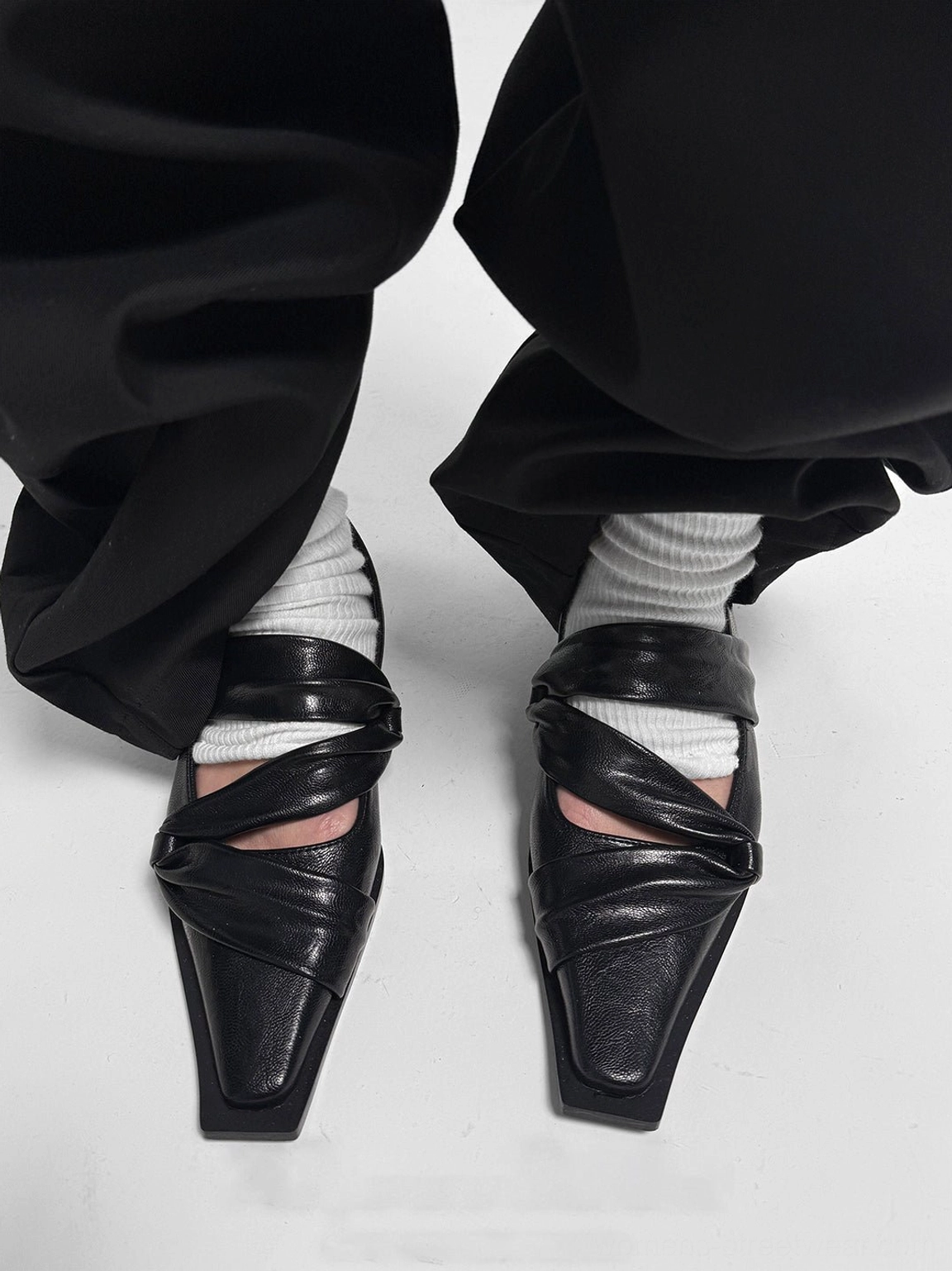 Front view of paired black knotted square-toe leather flats worn with white ribbed socks and relaxed black trousers