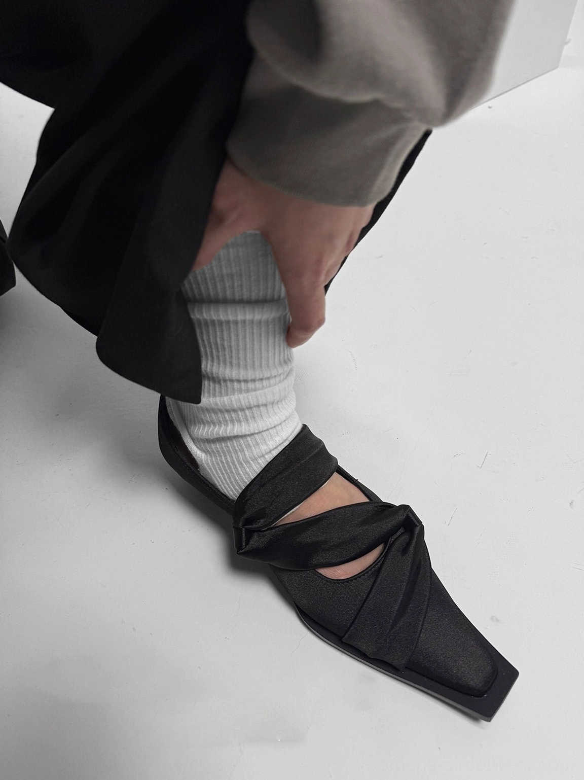 Black knotted leather flats worn with white socks, a grey top, and black trousers in a contemporary minimalist outfit