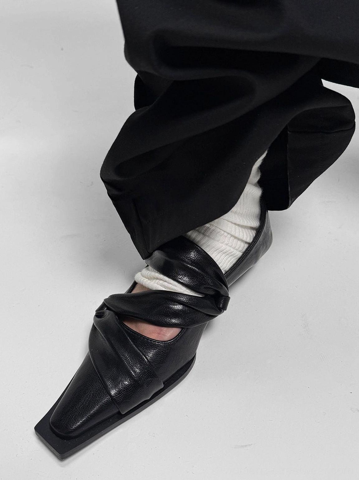 Low side view of black leather square-toe flats worn with white socks and wide-leg black pants for minimalist streetwear styling