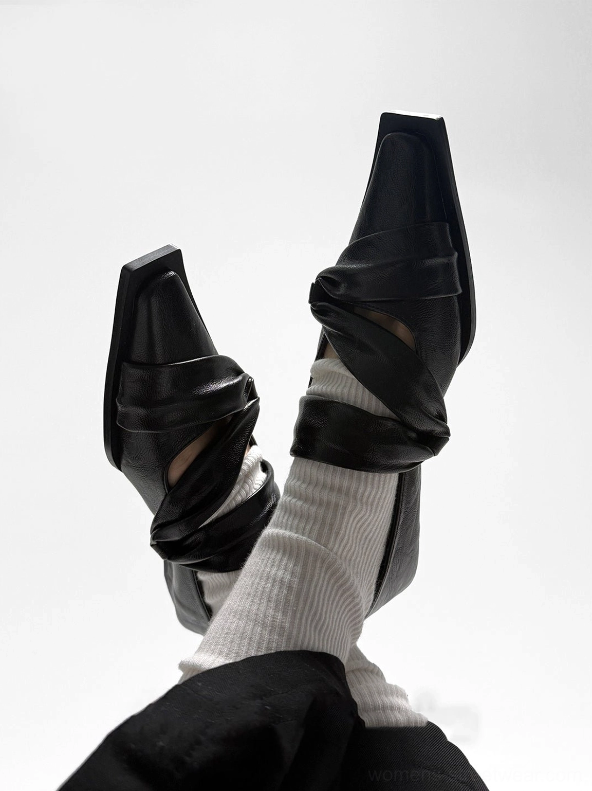 Overhead view of crossed feet wearing black knotted leather flats with twisted wrap straps and sharp geometric square toes