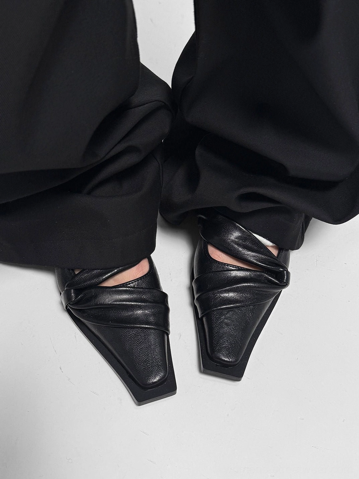 Top-down view of black square-toe leather flats with twisted cross straps styled under wide-leg black trousers