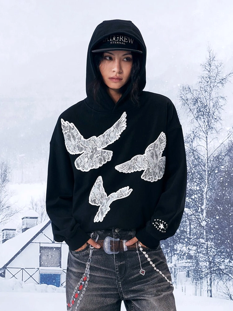 Front full-body view of a black lace dove statement hoodie styled with vintage jeans and chain accessories in a snowy winter streetwear setting