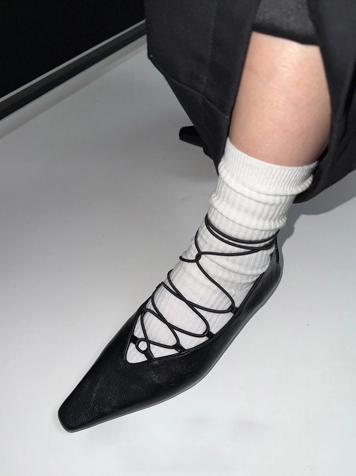 Side view of black lace-up ballet flat worn with white socks, featuring ankle-length tying laces, pointed square toe, and low block heel