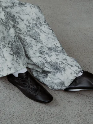Black lace-up ballet sneakers worn with white socks and printed loose trousers, showing the rounded toe and minimalist low-profile rubber sole