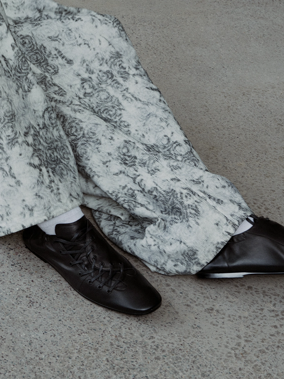 Black lace-up ballet sneakers worn with white socks and printed loose trousers, showing the rounded toe and minimalist low-profile rubber sole