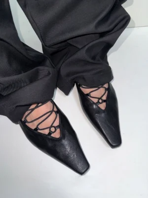 Black lace-up ballet flats worn with loose tailored black trousers, showing crossed instep straps, pointed square toe, and low heel