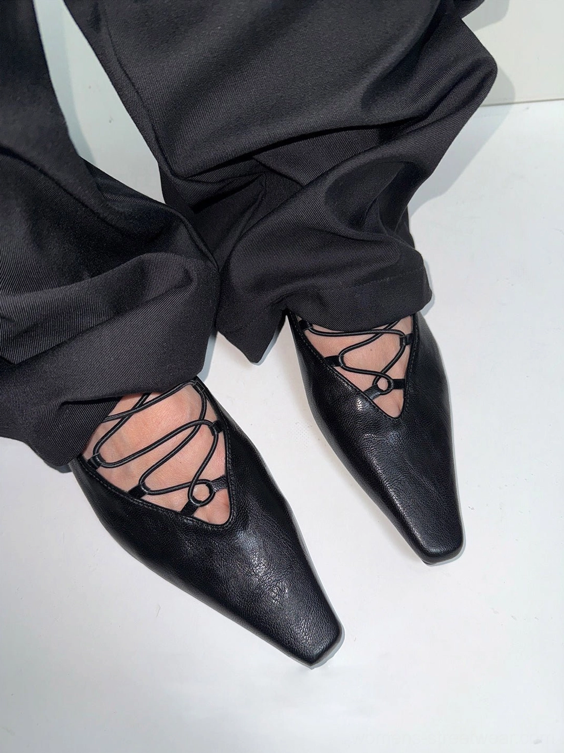 Black lace-up ballet flats worn with loose tailored black trousers, showing crossed instep straps, pointed square toe, and low heel