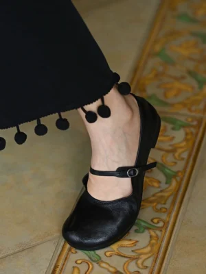 Black leather Mary Jane flat worn with cropped black streetwear trousers featuring pom-pom trim and a low flat sole