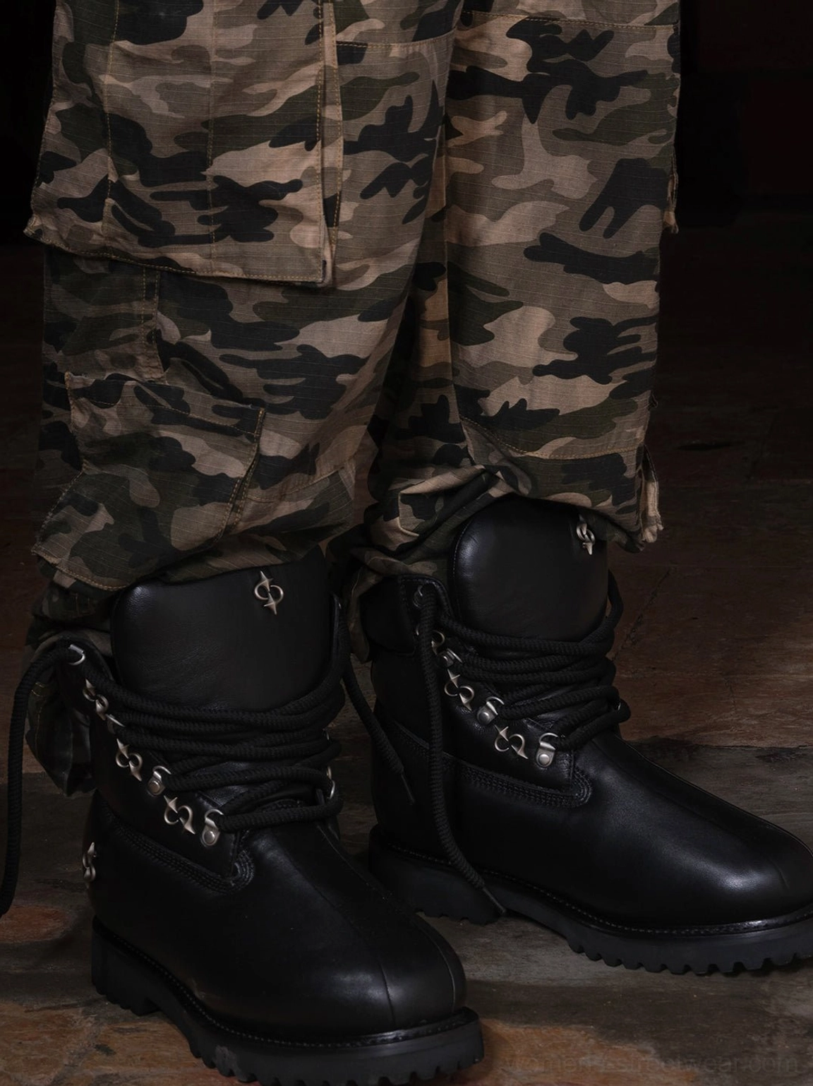 Black leather shearling work boots worn with camouflage cargo pants, showing the chunky ankle silhouette, fleece collar detail, utility styling, and thick lug sole