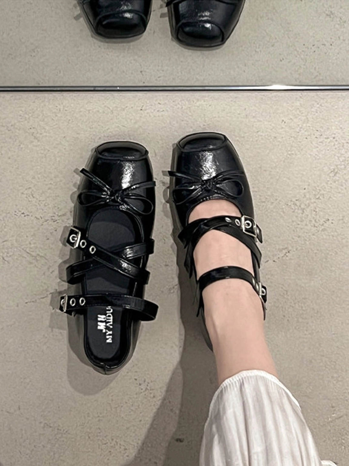 On-foot view of black leather square-toe Mary Jane flats with bow detail and white pleated skirt styling