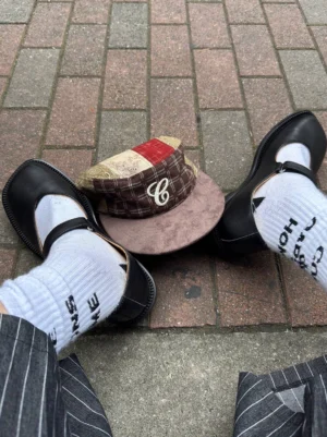 Black leather Mary Jane flats styled with white logo socks, pinstripe trousers, and a cap for a Seoul streetwear outfit on brick pavement