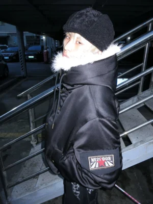 Side rear view of a black padded fur-collar bomber jacket with oversized hood and utility-inspired sleeve patch in an urban night setting