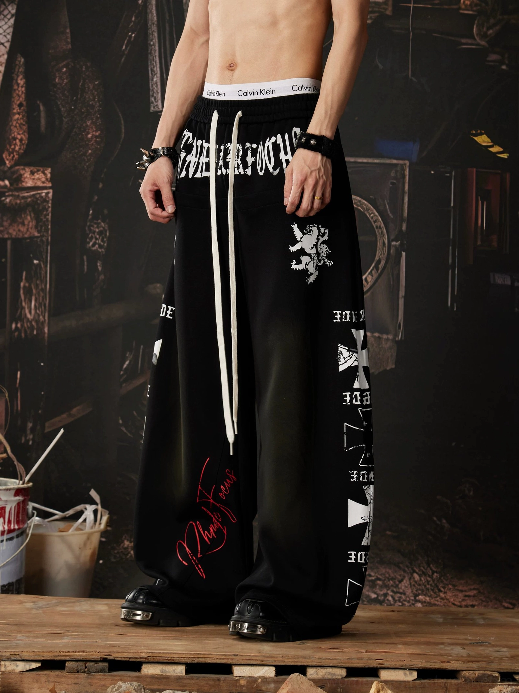 Black graphic wide leg streetwear sweatpants with large front lettering, heraldic lion artwork, cross side graphics, and elastic drawstring waistband