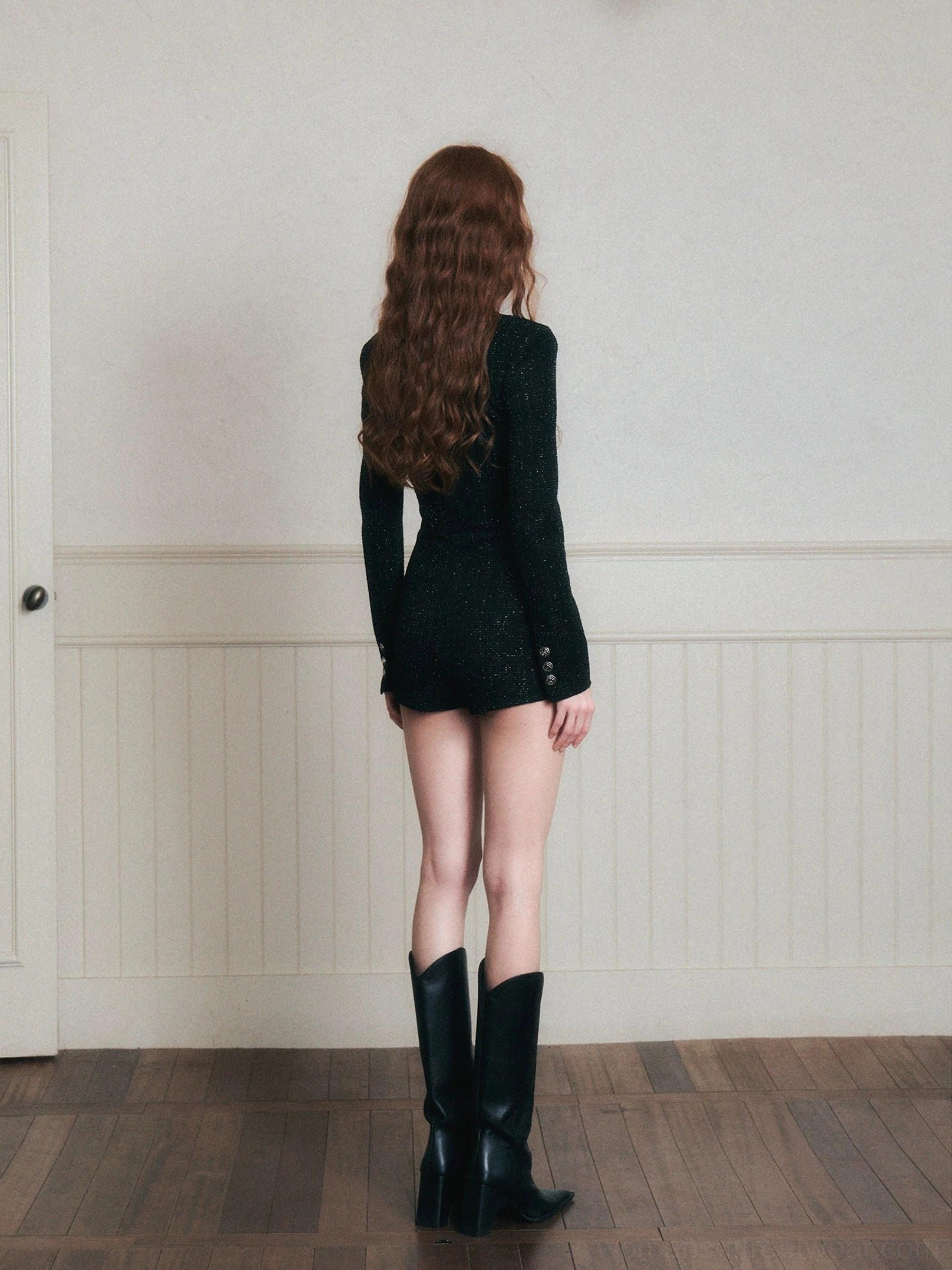 Back full-body view of a black long-sleeve tweed romper showing the fitted back, buttoned cuffs, tailored shorts, and sleek short playsuit silhouette