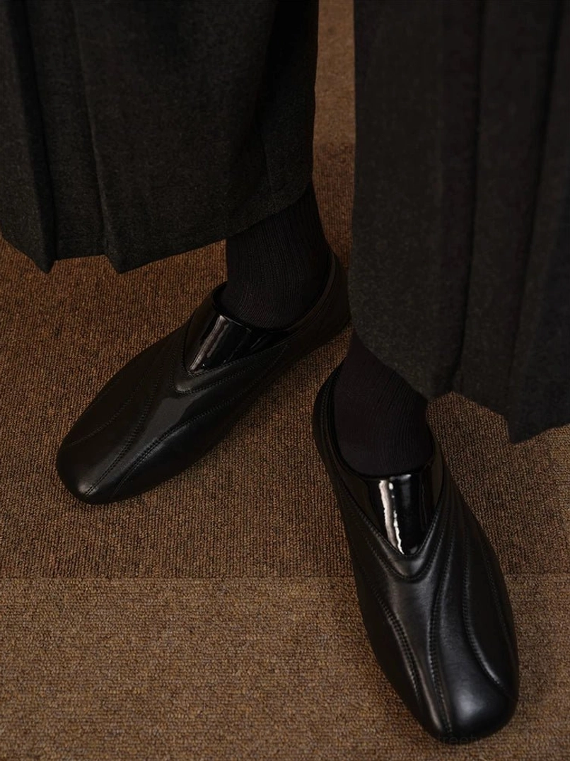 Side on-foot view of black low-profile leather slip-on shoes styled with black socks and a long black skirt