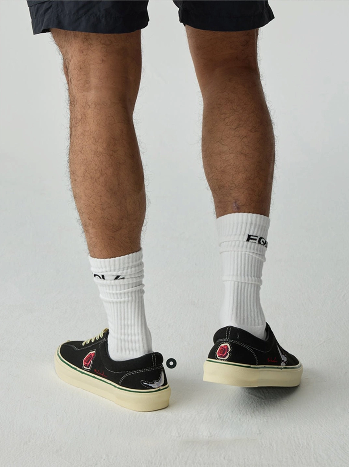 Rear view of black low-top skate shoes with classic off-white rubber soles worn with shorts and streetwear socks