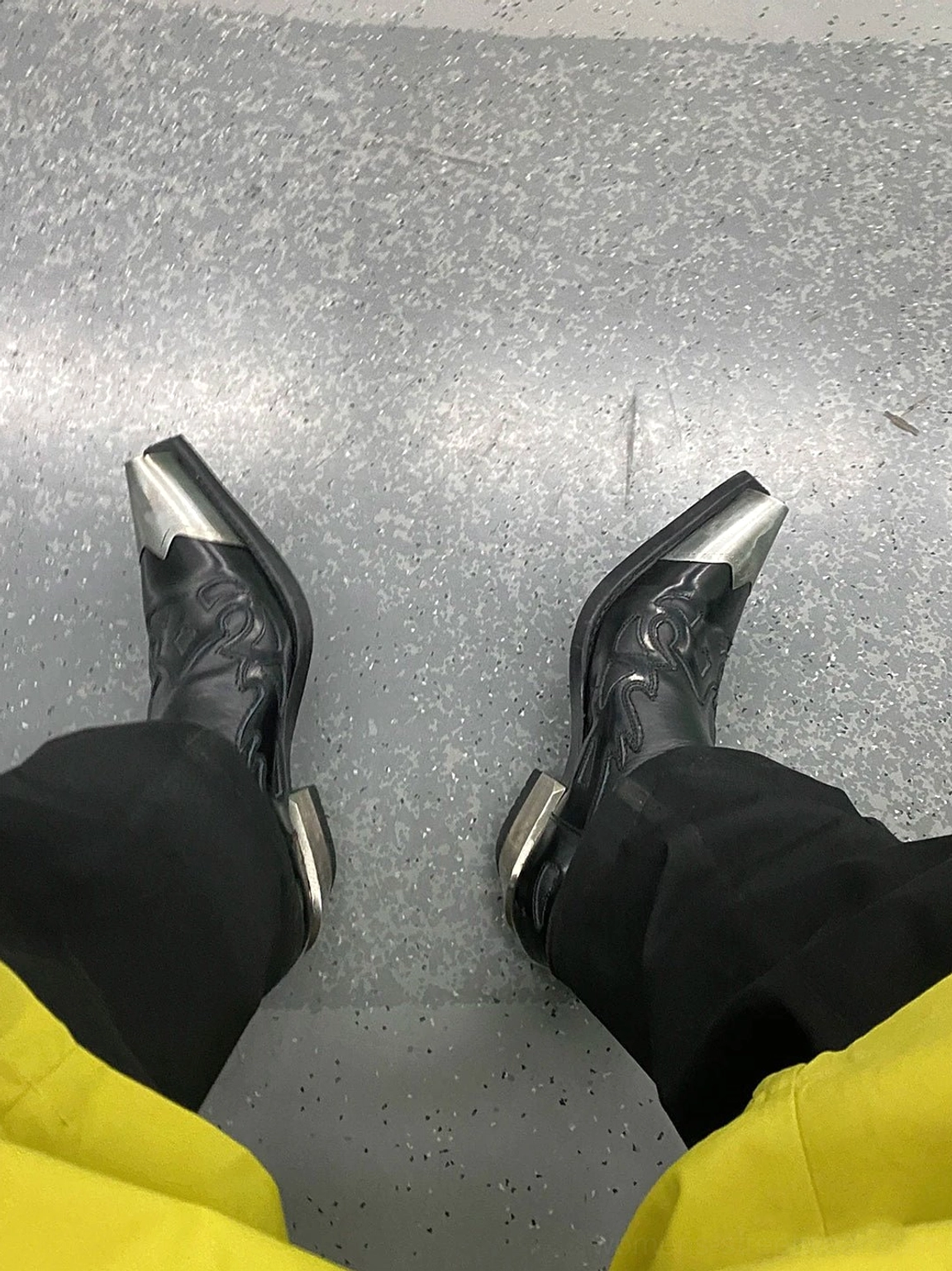 Top down on-foot view of black metallic toe block heel Chelsea boots styled with black trousers and yellow cropped pants on a concrete floor