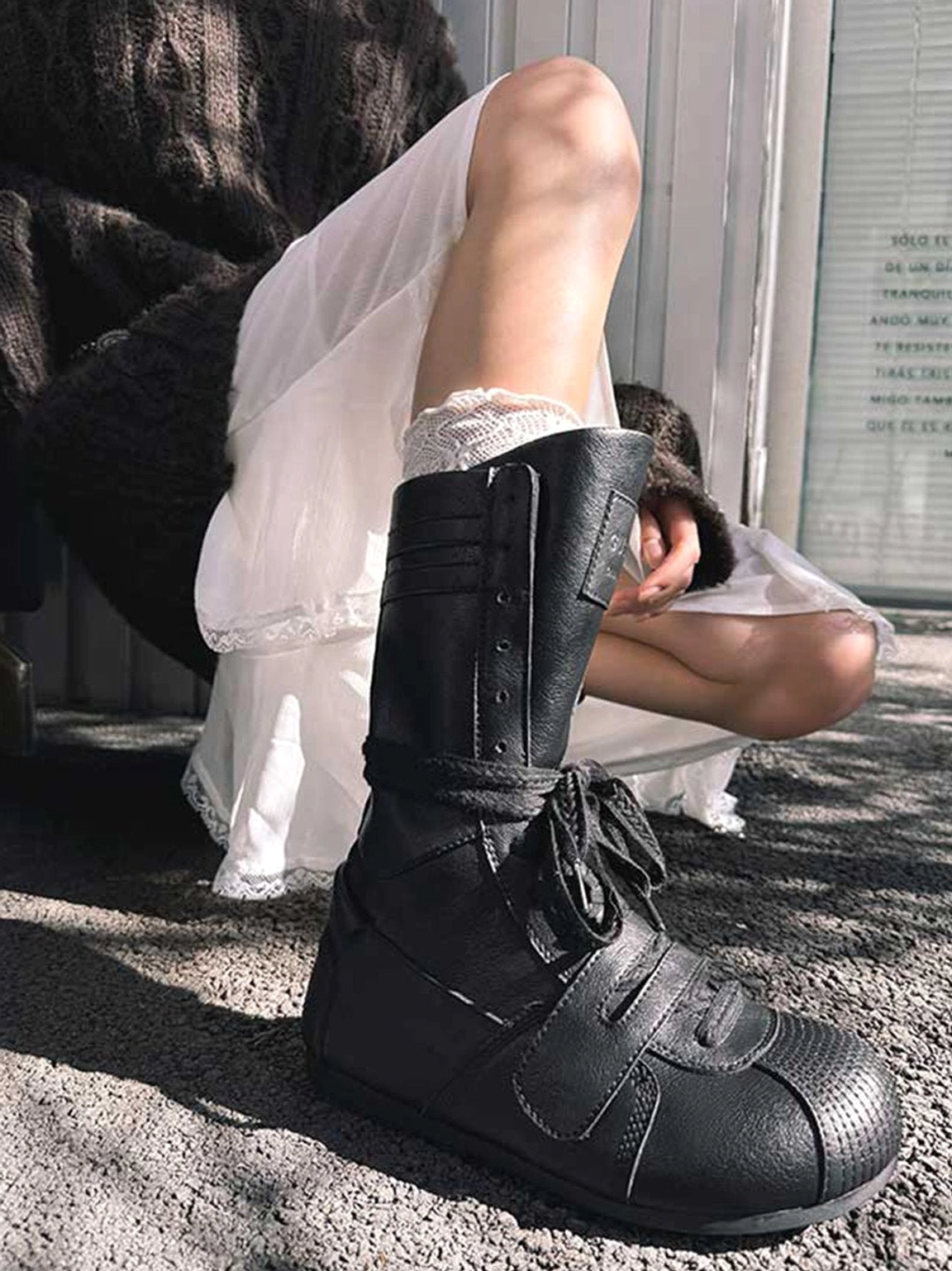 Side view of black leather-look mid-calf boxing boots styled with white lace socks, a sheer dress, and dark knitwear for avant-garde street fashion