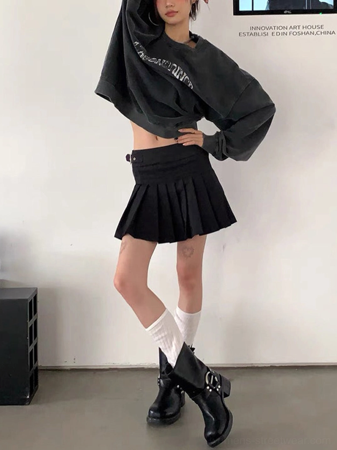 Side view of black mid-calf buckled leather cowboy boots styled with a pleated skirt and casual streetwear outfit
