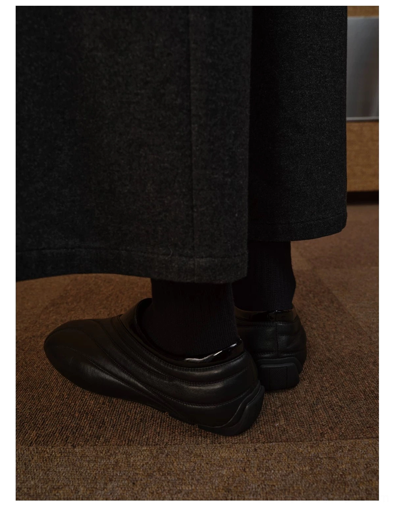 Rear on-foot view of black minimalist leather slip-on shoes with flat rubber sole and streamlined streetwear silhouette