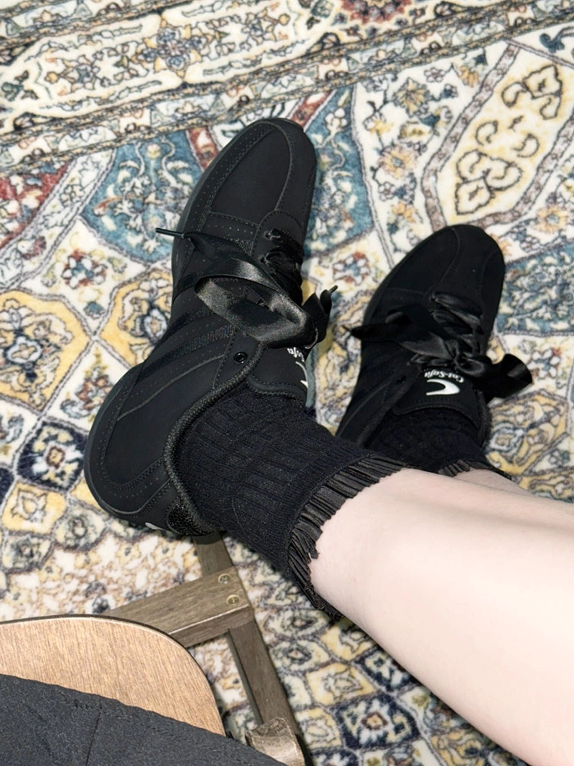 Top-down view of black minimalist ribbon lace sneakers with ruffled ankle socks on a vintage patterned carpet