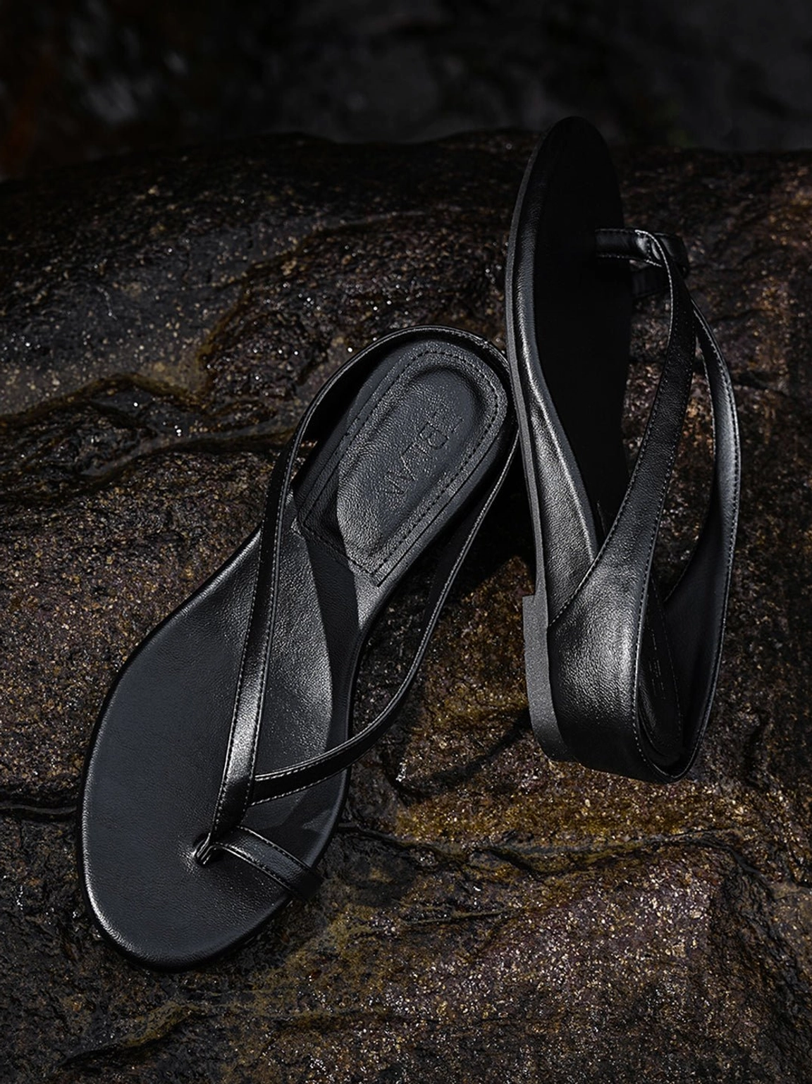 Black toe ring low wedge flip-flop sandal with visible footbed displayed on textured dark rocks