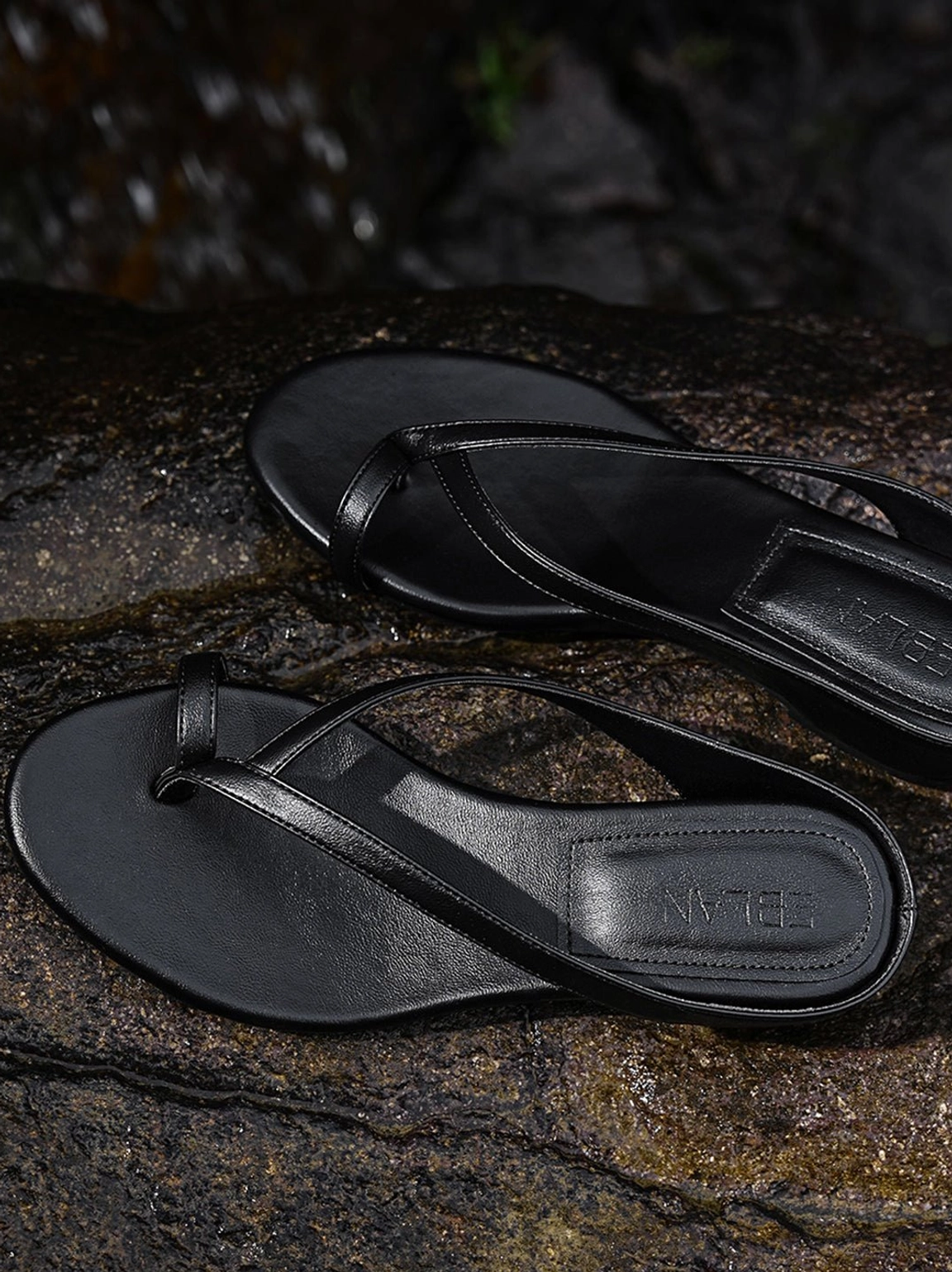 Top view of black minimalist toe ring wedge flip-flop sandals displayed on wet dark rocks