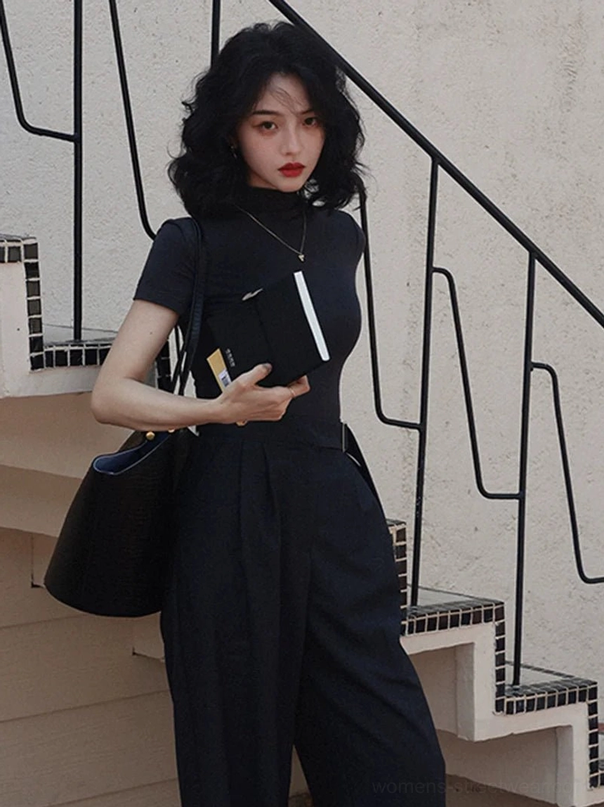 Woman wearing a black mock neck short sleeve slim fit streetwear top with high waisted dark wide leg trousers and tote bag on an outdoor staircase