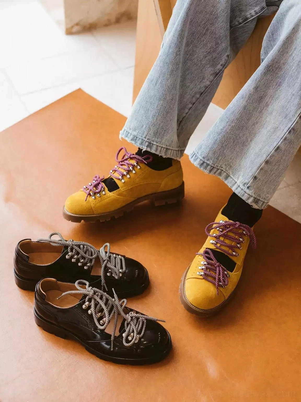 Two pairs of chunky streetwear derby shoes in black patent-look and mustard yellow suede-look finishes with braided laces on a wooden tabletop