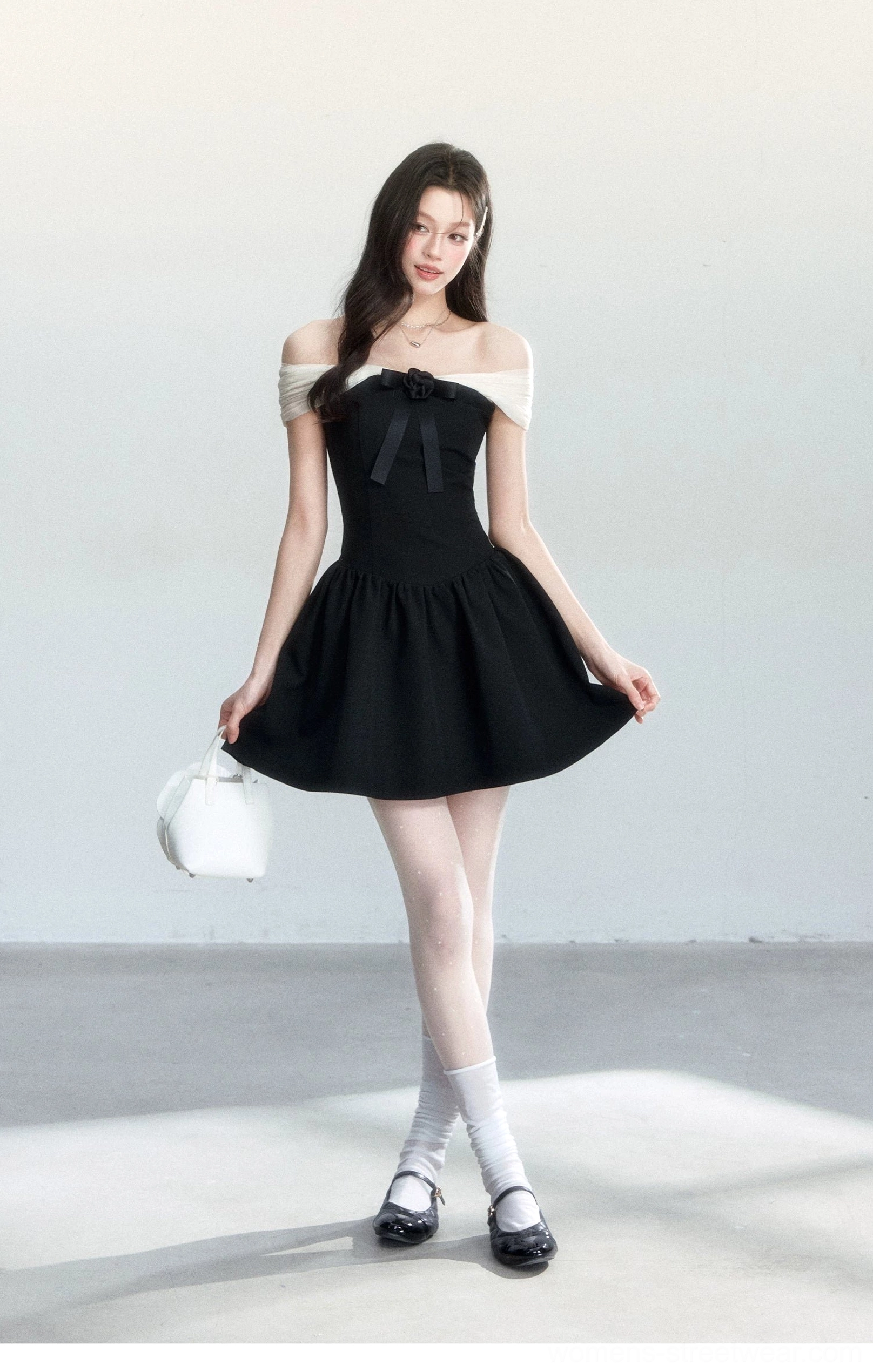 Side view of a black off-shoulder A-line mini dress with front bow, contrast neckline, white handbag, socks, and leather shoes