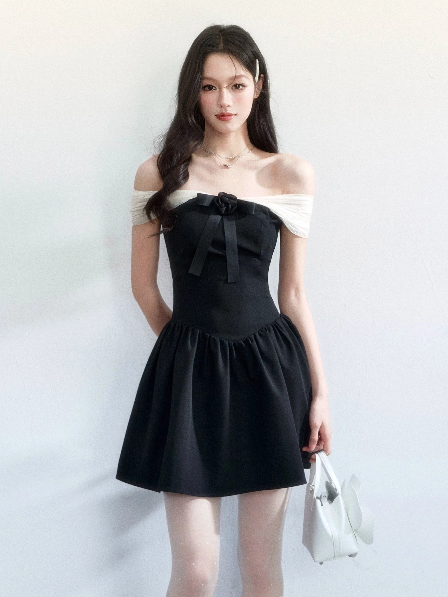 Back-side full-body view of a black off-shoulder bow mini dress with contrast trim, flared skirt, and white flower handbag