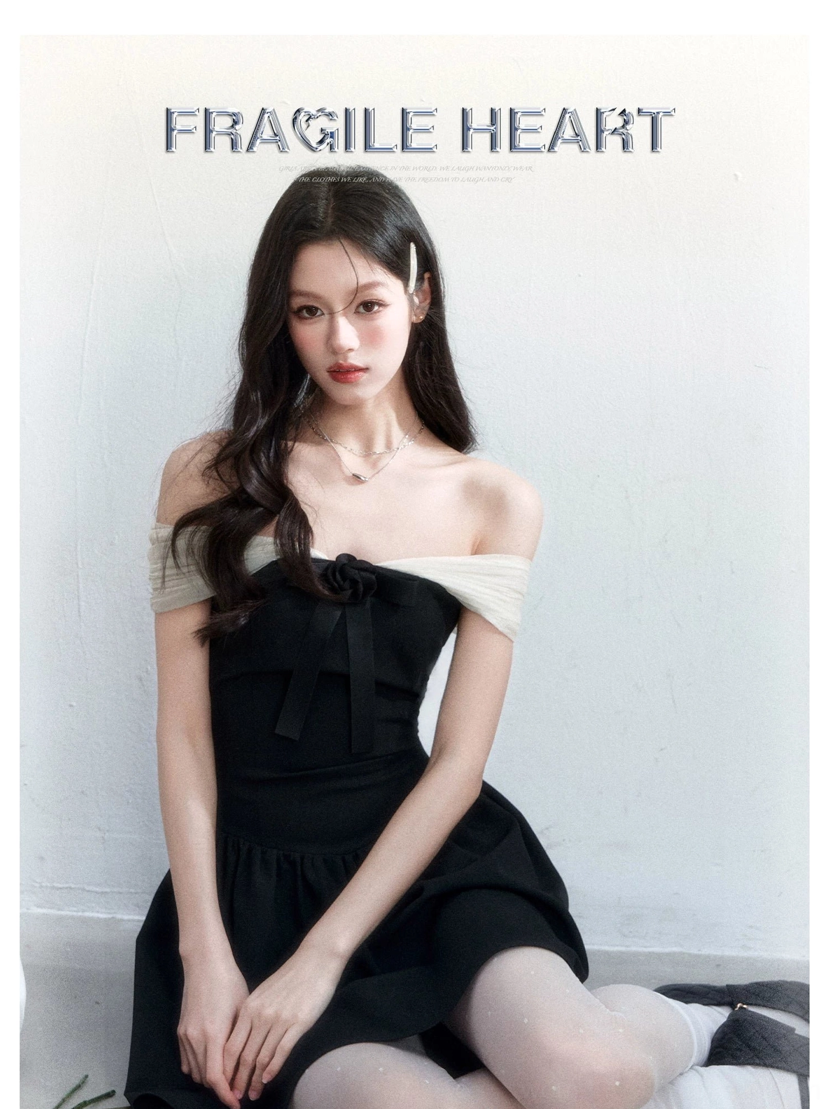 Black off-shoulder bow mini dress with light contrast mesh straps, slim waist, and flared skirt shown in a seated studio look