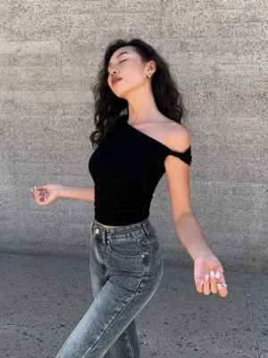 Black one-shoulder crop top shown in side profile with a diagonal neckline and high-waisted blue jeans