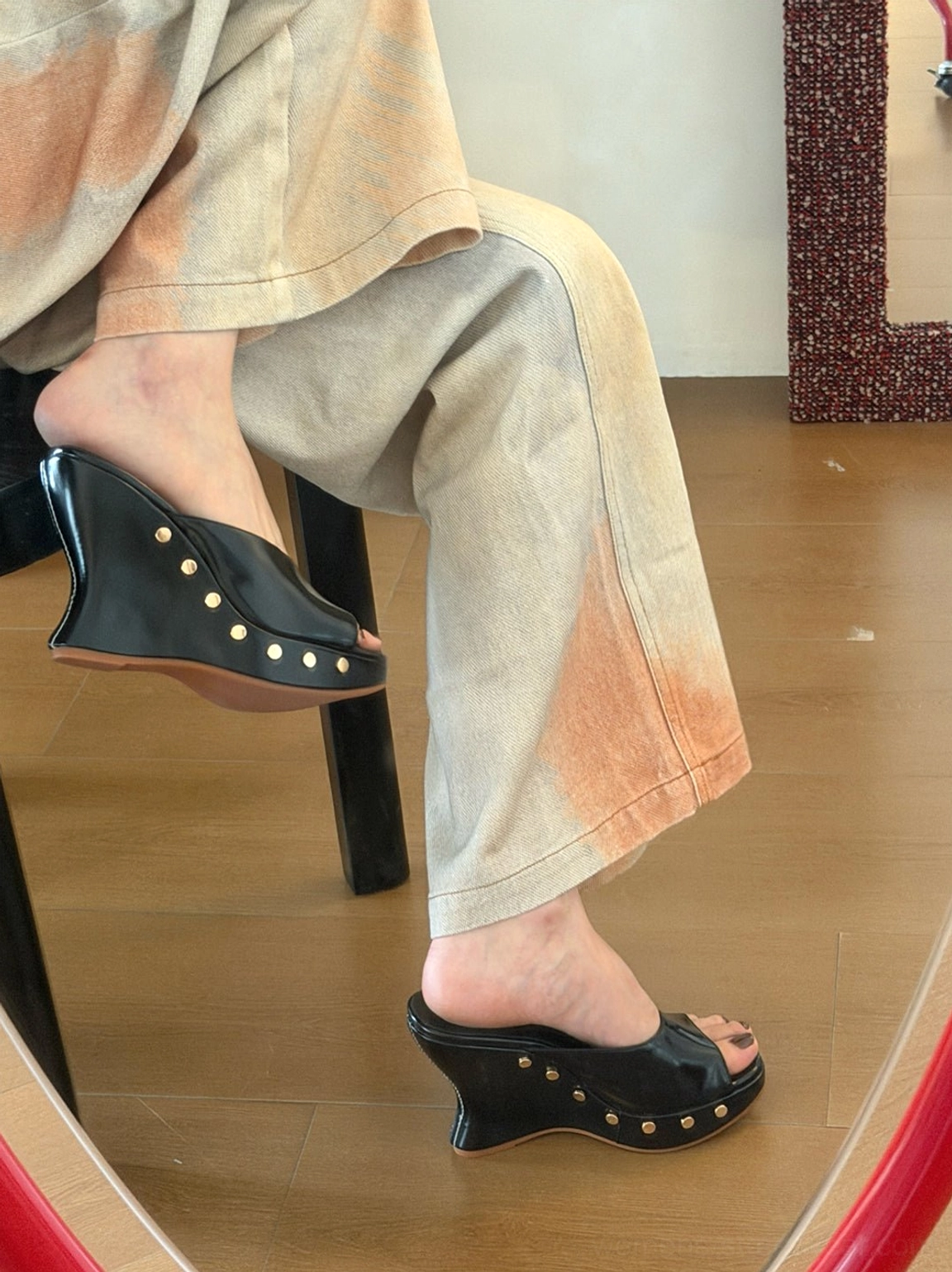 Rear view of black open-toe wedge mules worn with wide-leg streetwear trousers, showing brass-tone studs across the back of the sculpted wedge heel