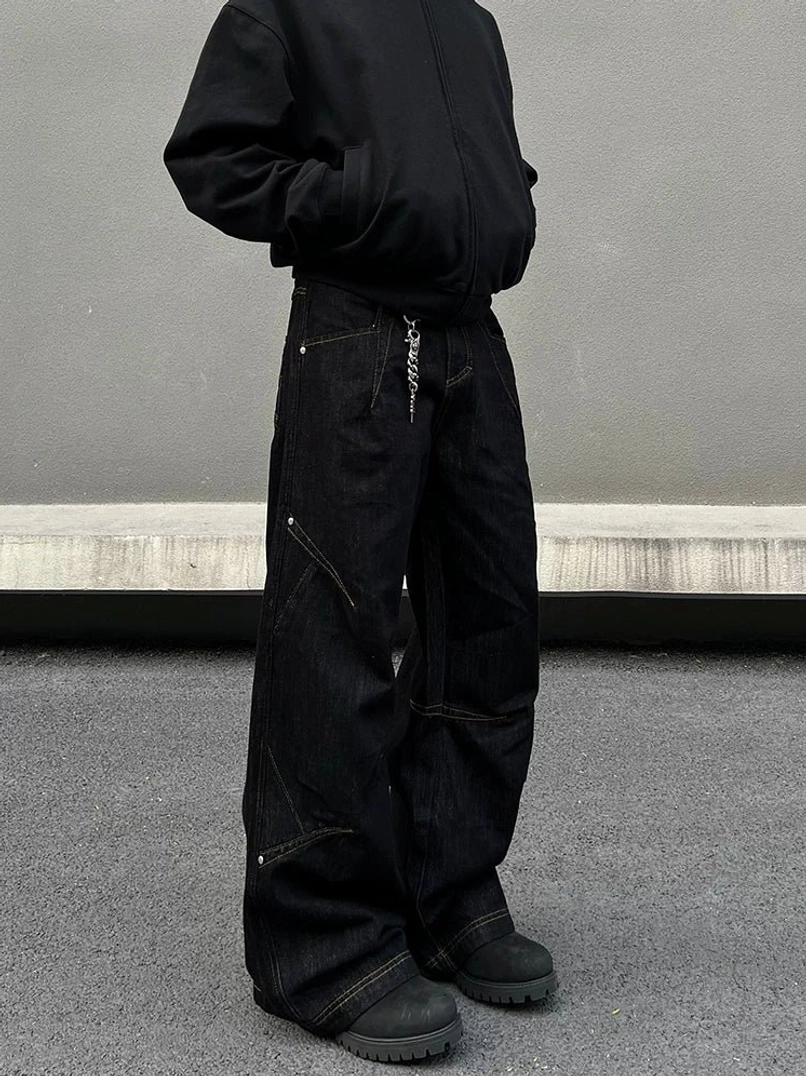 Front three-quarter view of oversized black wide-leg denim jeans featuring geometric panel seams, metal rivets, and contrast topstitching